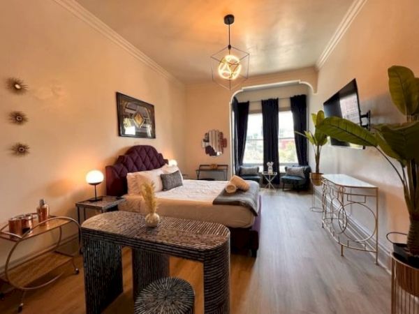 A stylish bedroom with a cozy bed, modern decor, potted plant, and soft lighting, featuring a large window with dark curtains.