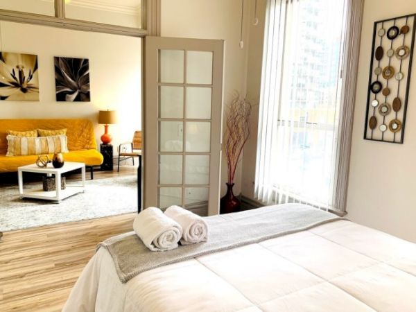 A bedroom with a made bed, rolled towels, and decorative art on the walls, connected to a living room with a sofa and modern decor.