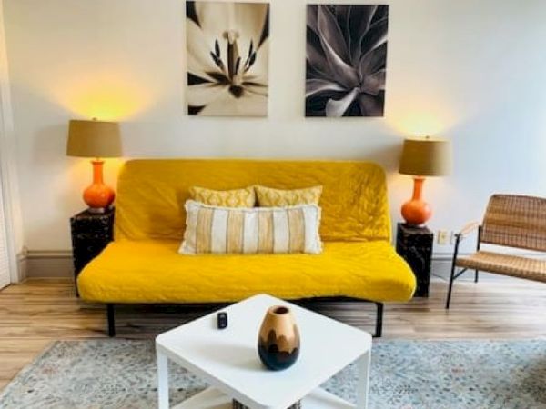 A cozy living room with a yellow sofa, cushions, vibrant lamps, wall art, and a small table with a decorated vase.