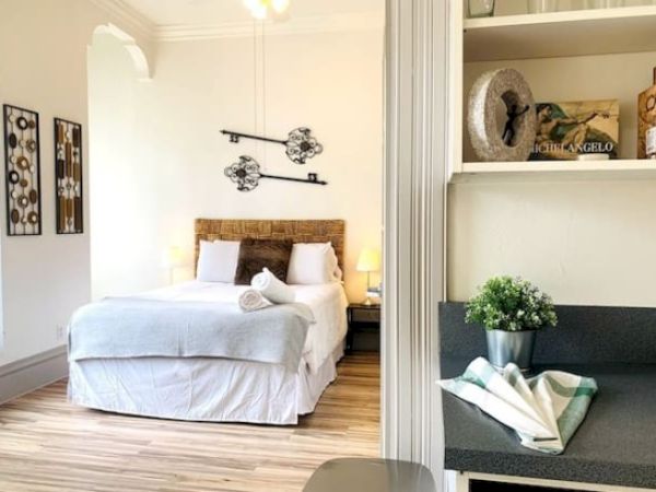 The image shows a cozy bedroom with a double bed, stylish decor, and a small kitchenette area.
