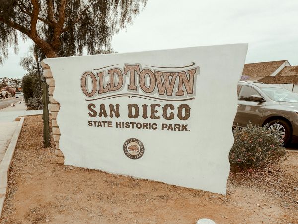 A sign for Old Town San Diego State Historic Park is displayed, with a car and trees in the background.