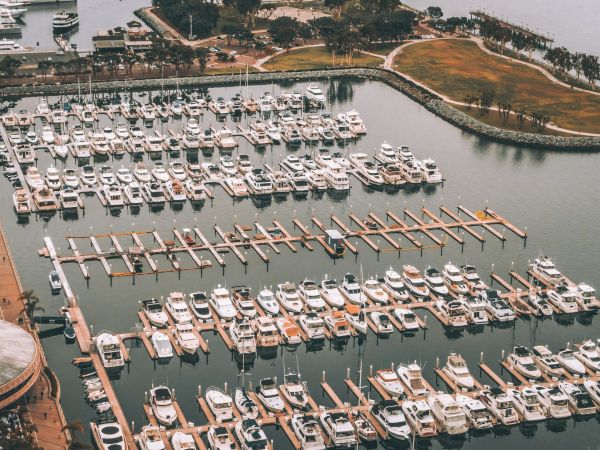 Aerial view of a marina with numerous boats docked, surrounded by water and a grassy area with trees in the background.