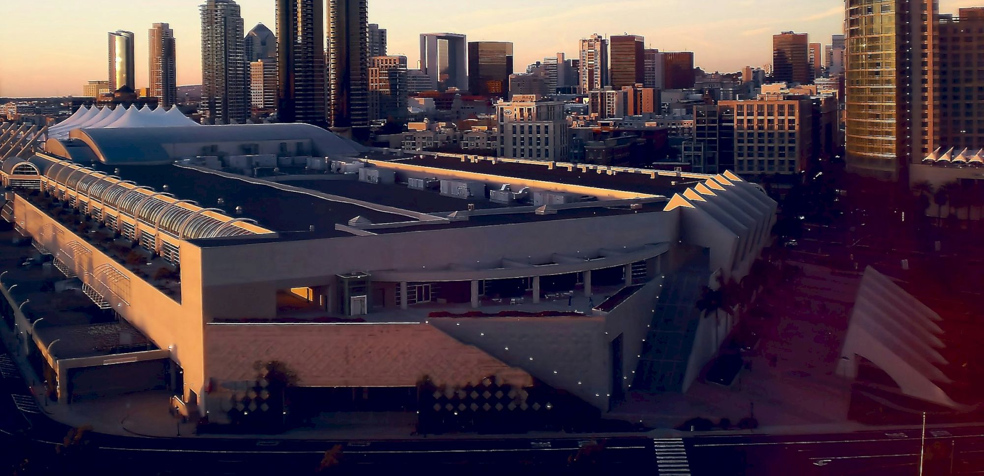 A large convention center with a modern design is surrounded by a cityscape with tall buildings during sunset.