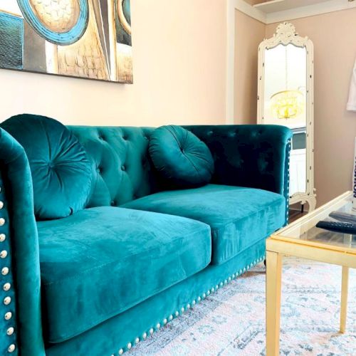 A teal couch with round cushions, a fashion magazine on a glass table, art on the wall, and a mirror in the room.