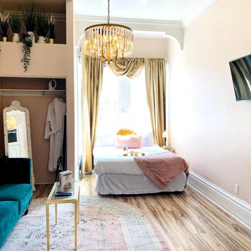 A stylish bedroom with a bed, green sofa, chandelier, plants, wall-mounted TV, and a mirror, creating a cozy, modern atmosphere.