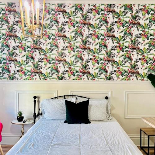 A bedroom features a bold jungle-themed wallpaper, a black iron bed, bright pink chair, plants, side table, and unique hanging light fixture.