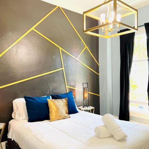 Modern bedroom with geometric wall design, white bedding, blue and gold pillows, and elegant lighting fixtures.