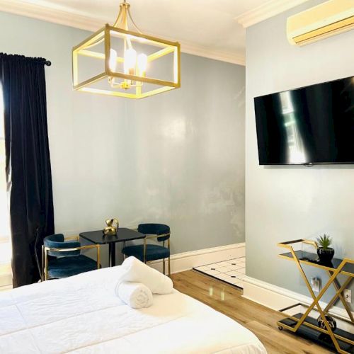 A modern bedroom with a bed, rolled towels, wall-mounted TV, small table, chairs, and air conditioner. Black curtains and gold accents.