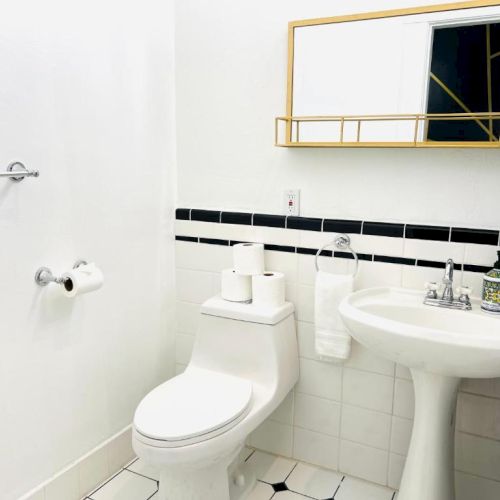 This image shows a bathroom with a toilet, pedestal sink, mirror, towel rack, toilet paper, and soap dispenser, all in a white and black theme.
