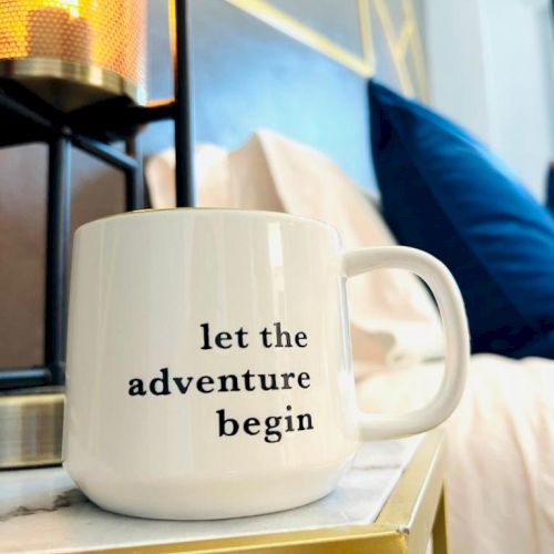 A white mug with "let the adventure begin" printed on it, placed next to a lamp. There are pillows and a bed in the background.