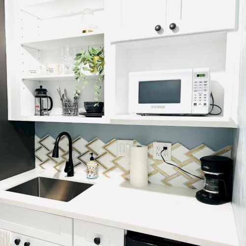 A kitchen corner with a sink, microwave, coffee maker, cups, plants, and black faucet on a white countertop with geometric backsplash.