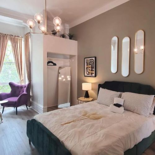 A cozy bedroom with a bed, purple chairs, lamps, and abstract art on the walls, featuring a large window with drapes.