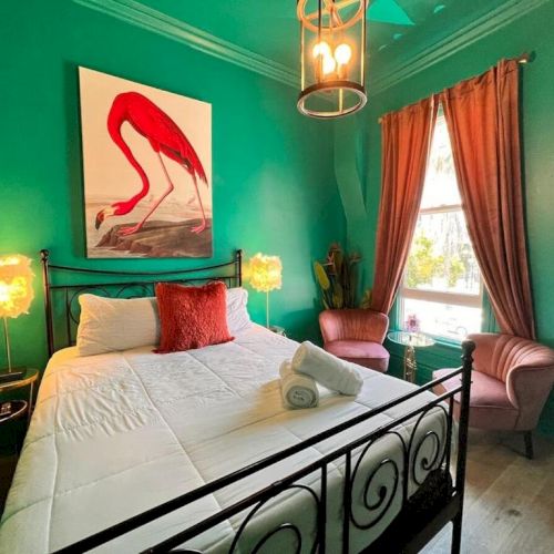 A vibrant bedroom with teal walls, a flamingo painting, a bed with red accents, two pink chairs, and warm lighting from unique lamps.