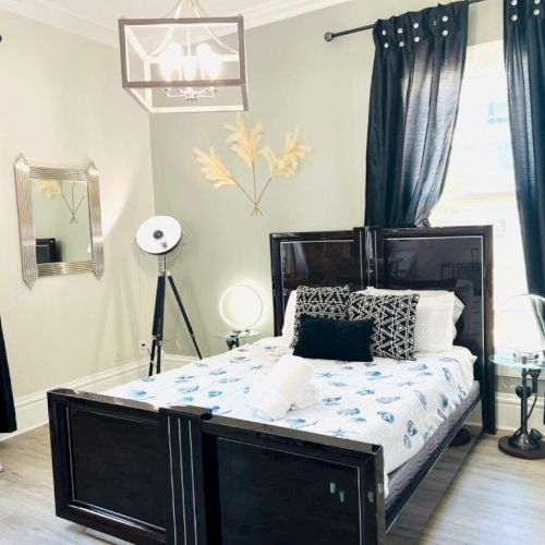 A modern bedroom with a dark wood bed, dark curtains, decorative pillows, a tripod lamp, a wall mirror, and a unique ceiling light fixture.
