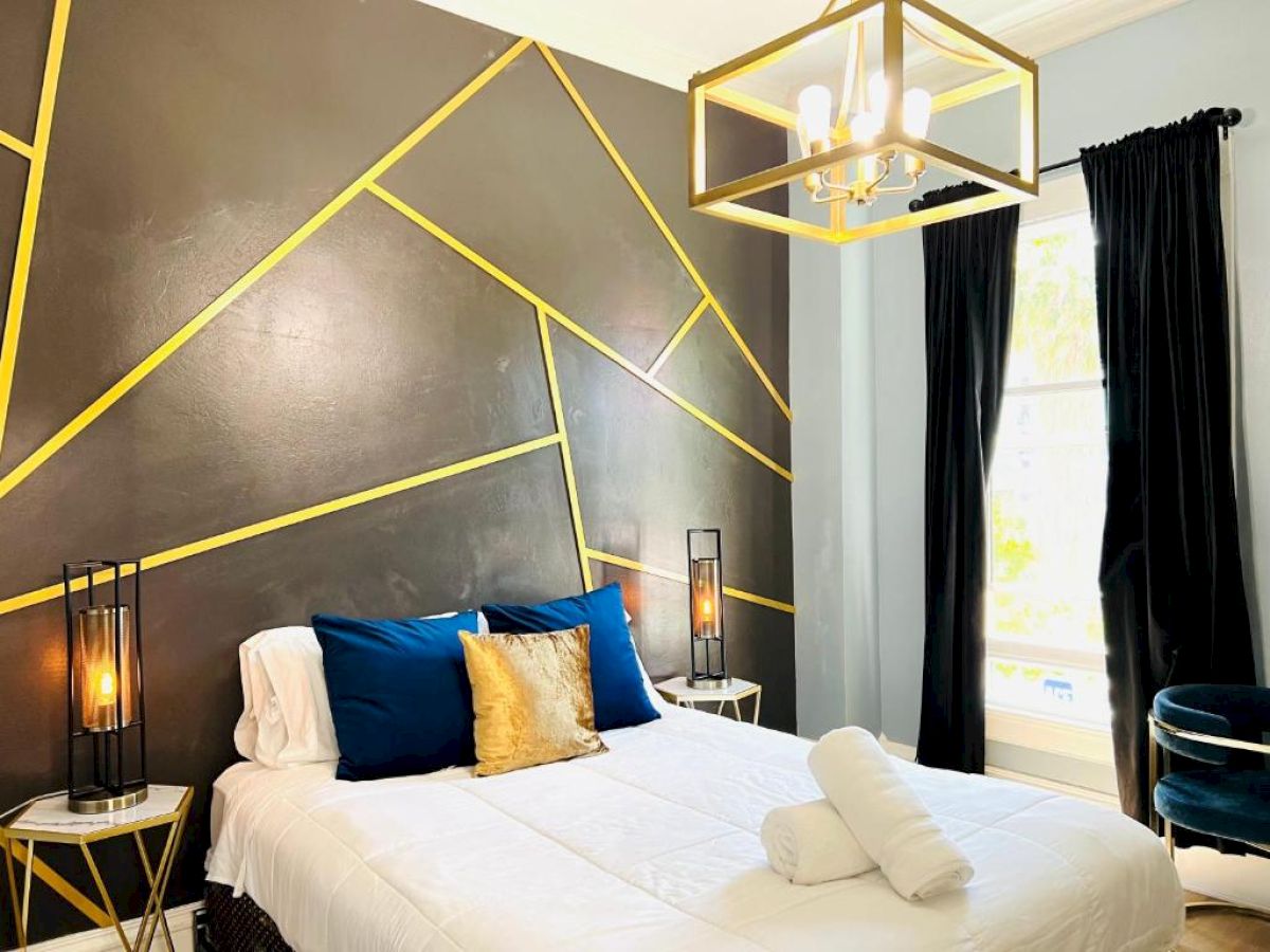 A modern bedroom with geometric black and gold wall, bed with blue and gold pillows, rolled towels, and hanging light fixture.