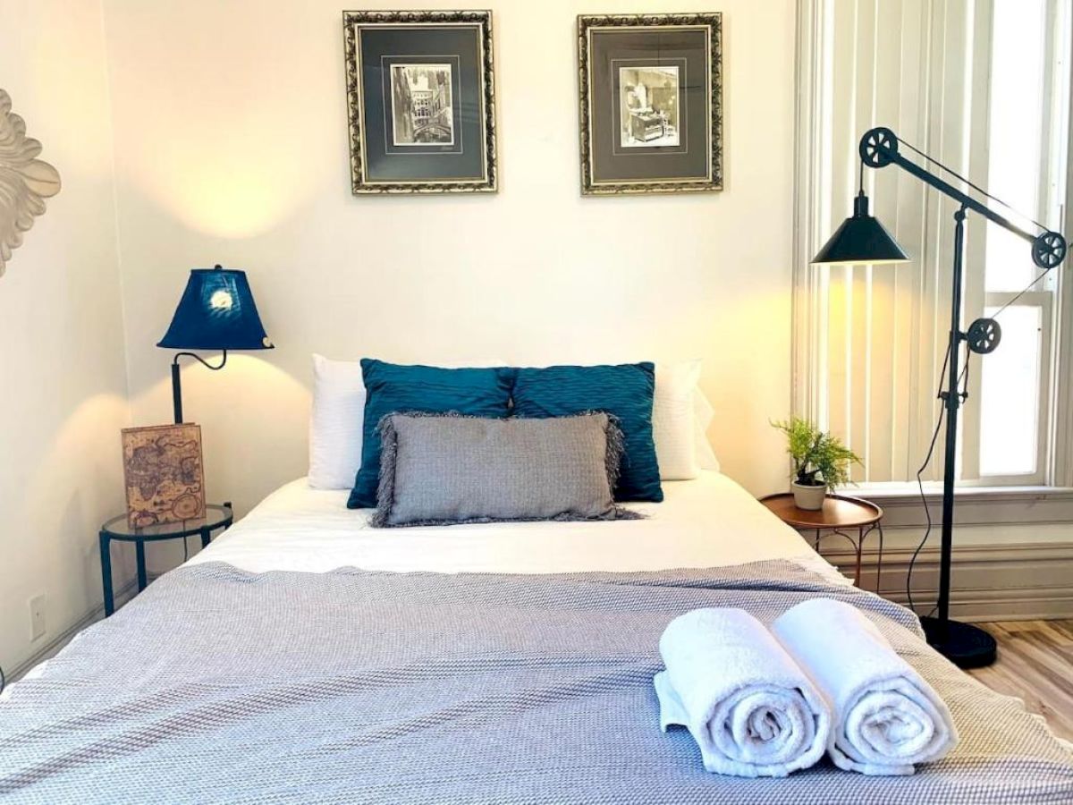 A stylish bedroom with a neatly made bed, two framed pictures, bedside lamps, and rolled towels at the foot of the bed.
