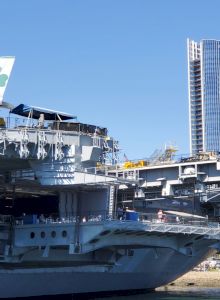 An aircraft carrier is docked by the water with a plane on deck, and modern city buildings in the background.