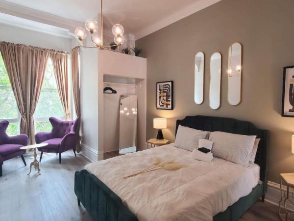 A stylish bedroom with a bed, wall mirrors, and modern decor. Purple chairs and a small table are near the window with light curtains.